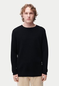 Pull noir texturé avec motif côtelé, encolure ronde et manches longues. Le matériau semble doux, avec une coupe décontractée.