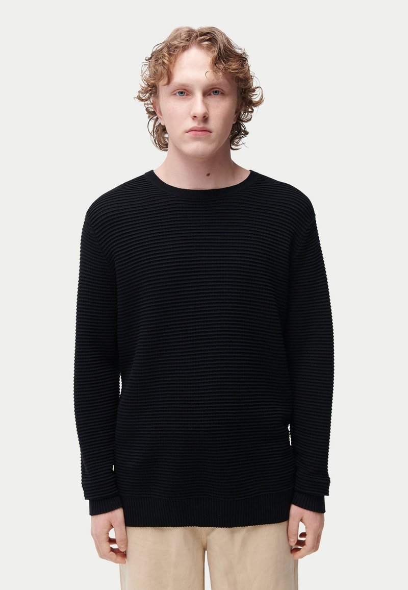 Pull noir texturé avec motif côtelé, encolure ronde et manches longues. Le matériau semble doux, avec une coupe décontractée.