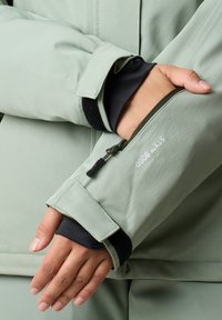 Light green outdoor jacket sleeve with zippered pocket, black inner cuffs, and Velcro fastening; features waterproof text detail.