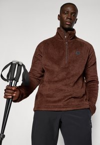 Brauner Fleece-Pullover mit Reißverschluss aus strukturiertem Stoff. Mit einem Logopatch und hohem Kragen. Gehalten mit Trekkingstöcken in schwarzen Hosen.