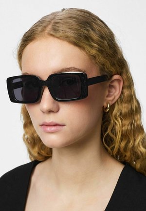 Mladá osoba s vlnitými blond vlasmi, ktorá má na sebe veľké čierne obdĺžnikové slnečné okuliare a malé zlaté náušnice v tvare kruhu na jednoliatom pozadí.