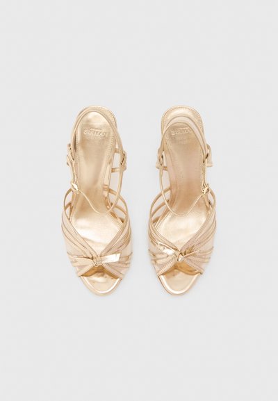 Χρυσά μεταλλικά τακούνια με ανοιχτά δάχτυλα και λουράκια που διασταυρώνονται, διακοσμημένα με μικρά φιόγκους σε απλό ανοιχτό γκρι φόντο.