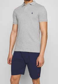 Man som bär en grå kortärmad polo och marinblå shorts, stående mot en enkel bakgrund.