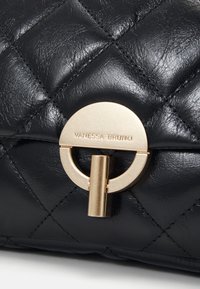 Čierna prešívaná kožená kabelka s pozlátenou kruhovou sponou a vertikálnou tyčou. Na sponke je embosovaný text "VANESSA BRUNO".