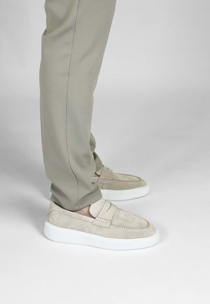 Beige suède instap schoenen met een witte zool, voorzien van stikseldetails en een ronde neus. G gedragen met lichtbruine, taps toelopende broek.
