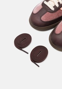 Bruna läderskor med rosa mockapaneler, krämfärgade skosnören och gummisulor; två matchande bruna snören ligger bredvid dem.