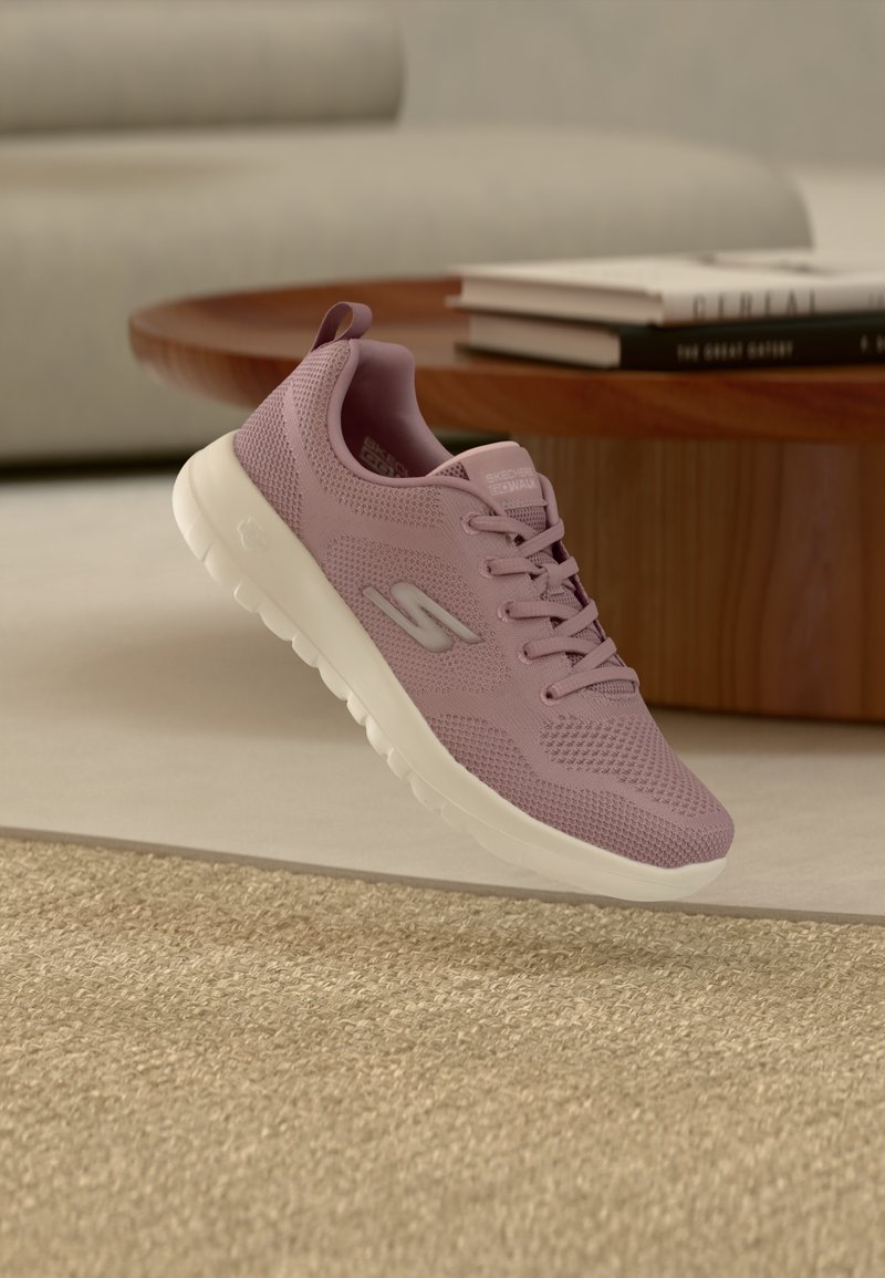 Zapatilla de punto color púrpura claro con suela blanca flota sobre una alfombra beige, mesa de café de madera y libros en un entorno de sala de estar de tonos neutros.