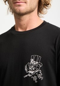 Man wearing black shirt with white graphic of a skull in a top hat smoking a cigar above two rolling dice on the chest.