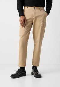 Beige cotton cargo pants with two side pockets, tapered legs, and a button closure, paired with black polished shoes and dark socks.