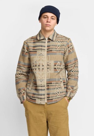 Beige zip-up jack met geometrische patronen in tan, bruin en grijs. Heeft een kraag en lange mouwen, gemaakt van textuurstof.