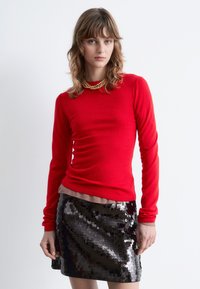 Red long-sleeve top with ribbed texture layered over a beige shirt, paired with a shiny black sequined mini skirt featuring a fitted design.