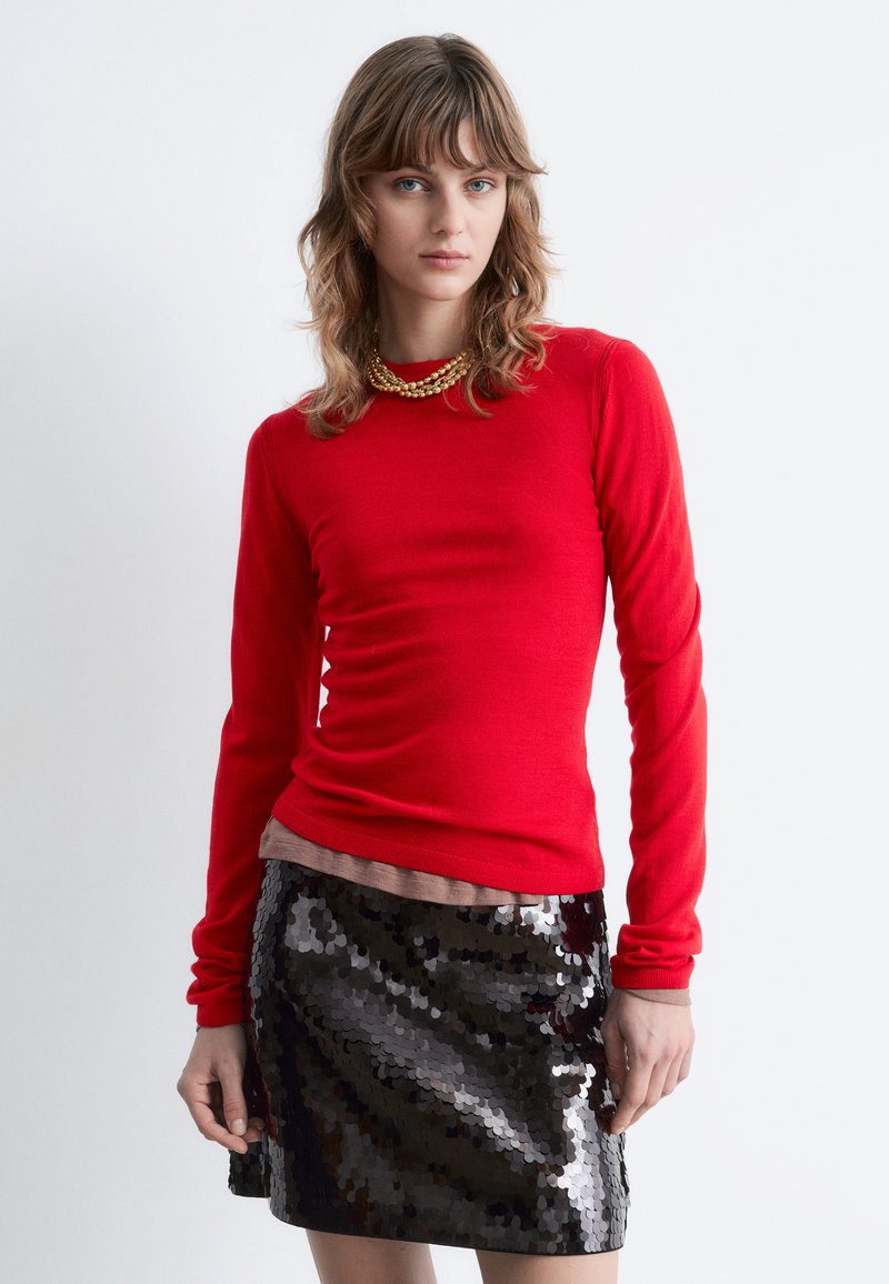 Red long-sleeve top with ribbed texture layered over a beige shirt, paired with a shiny black sequined mini skirt featuring a fitted design.