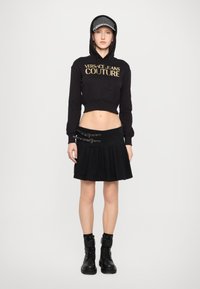Svart kort hoodie med "Versace Jeans Couture"-logotyp, i kombination med en svart plisserad kjol med spännen och svarta ankelstövlar.