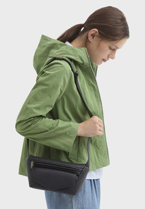 Jonge vrouw in een groene capuchonjas en blauwe spijkerbroek, die een zwarte leren crossbodytas met een ritsvak vasthoudt.
