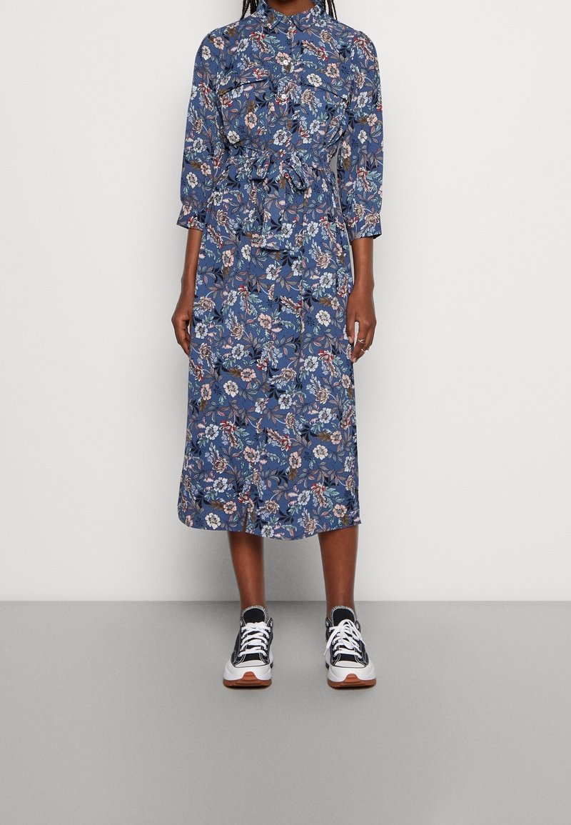 Robe midi bleue à motif floral, manches longues, taille nouée et détails à boutons, portée avec des baskets noires et blanches.