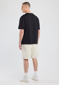 Camiseta negra de cuello redondo con mangas cortas, combinada con pantalones cortos beige. El conjunto se complementa con zapatillas blancas y calcetines tobilleros.
