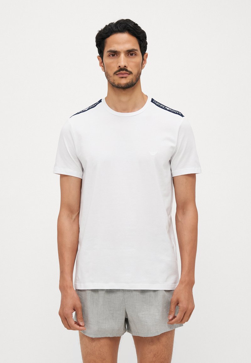 T-shirt bianco in cotone a maniche corte, con dettagli navy sulle spalle e un piccolo logo sul petto. Indossata con pantaloni corti grigi chiari.