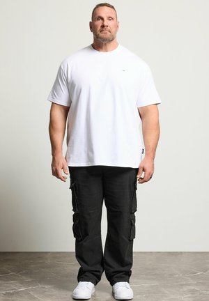 Uomo in piedi di fronte, indossa una maglietta bianca semplice, pantaloni cargo neri e scarpe da ginnastica bianche su un pavimento grigio.