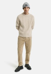 Suéter de lana beige de punto, pantalones de pana crema y botas negras con cordones. Gorro de punto gris con un pequeño logo. Look simple y casual.