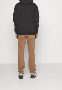 Schwarzer Hoodie, braune Hose und grünliche Schuhe. Der Hoodie hat eine Kängurutasche; die Hose ist gerade geschnitten und hat sichtbare Nähte.