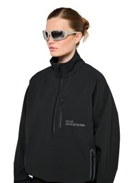 Frau trägt eine schwarze Jacke mit Reißverschluss und hohem Kragen sowie graue, umschließende Sonnenbrille, Haare zu einem Dutt gebunden.