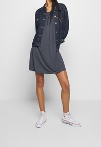 Šedé šaty s rozšířeným střihem, vrstvené pod tmavou riflovou bundou. Modelka nosí bílé vysoké tenisky a pózuje s jednou rukou v kapse bundy.