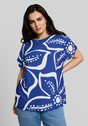 Frau mit langen dunklen Haaren, die eine blaue Bluse mit großem weißem Blumenmuster und hellblaue Jeans trägt, steht mit einer Hand in der Hüfte.