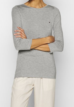 Person iført en grå langærmet top med en lille logo på brystet og lyse beige bukser, armene krydset ved livet.