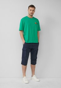 T-shirt en coton vert avec un logo, associé à un short cargo marine avec poches, et des baskets blanches. Design simple et décontracté.