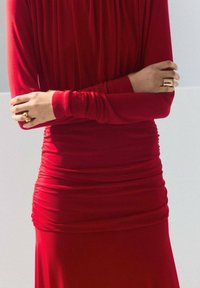 Rochie roșie, mulată, dintr-un material elastic, cu detalii strânse în talie și mâneci lungi; asortată cu inele aurii pe degete.