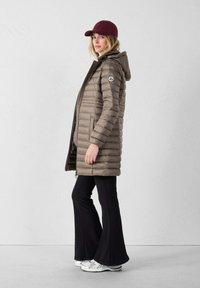 Vrouw in een lange taupe pufferjack, zwarte flared broeken, witte sneakers en een bordeauxrode baseballpet die zijwaarts tegen een plain muur staat.