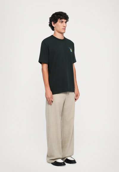 Tânăr îmbrăcat cu un tricou grafic negru, pantaloni largi bej și pantofi alb-negru, stând în fața unui fundal alb simplu.