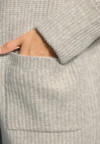 Cardigan lavorato a maglia grigio chiaro con textura a coste, dotato di una grande tasca anteriore e polsini ripiegati. Materiale morbido e accogliente.
