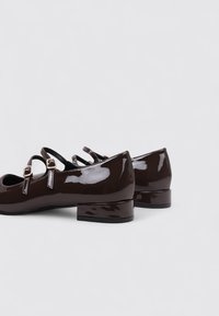 Zapatos de charol marrón con un bajo tacón bloque, que cuentan con dos correas ajustables y un diseño de punta redonda. Acabado liso y brillante.