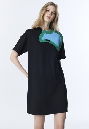 Jonge vrouw in een losse zwarte jurk met korte mouwen en asymmetrische blauwgroene gebreide patch op de schouder, staand met één hand op de heup.