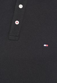 Svart texturerad polo skjorta med två vita knappar, en sydd med röd tråd, samt en liten rektangulär logotyp i rött, vitt och blått.