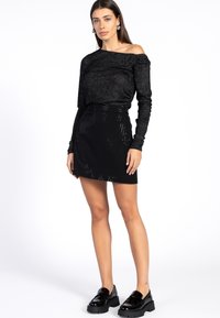 Zwarte off-shoulder top met lange mouwen in een gestructureerde stof, gecombineerd met een zwarte mini-jurk met een ribbelpatroon en sprankelende accenten. Zwarte plateau-schoenen.
