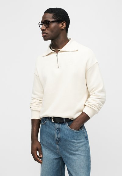 Άνδρας που φοράει φαρδιά κρεμ μπλούζα με γιακά φερμουάρ, μπλε τζιν, μαύρη ζώνη, κολιέ με πέρλες και σκοτεινά γυαλιά ηλίου, στέκεται με το ένα χέρι στην τσέπη.