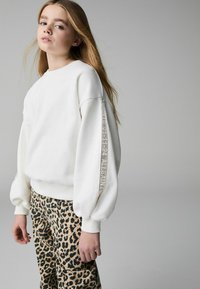 Vit sweatshirt med ribbade muddar och texten "ALLSAINT'S 01-11-94" på ärmen, matchad med leopardmönstrade byxor.