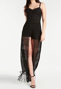 Schwarzes Häkelkleid mit offenem Vorderteil, das über komplexe Muster, einen figurbetonten Ausschnitt und Fransen am Saum verfügt. Getragen über Shorts und Sandalen.