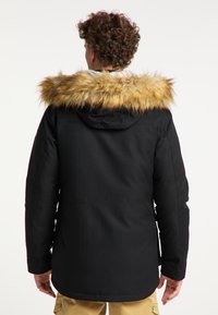 Mężczyzna w czarnej zimowej kurtce z kapturem i brązowym futrzanym obszyciem, sfotografowany od tyłu na jednolitym tle.