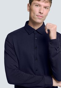 Diepblauw overhemd met lange mouwen, een klassieke kraag, knoopsluiting en een gestructureerde stof. Heeft een subtiele visgraatstructuur en manchetten.
