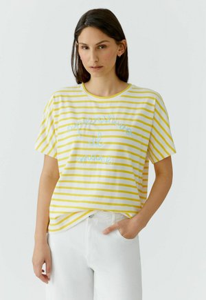 Vrouw die een geel-wit gestreept T-shirt draagt met geborduurde tekst en een witte broek, staand tegen een effen lichte achtergrond.