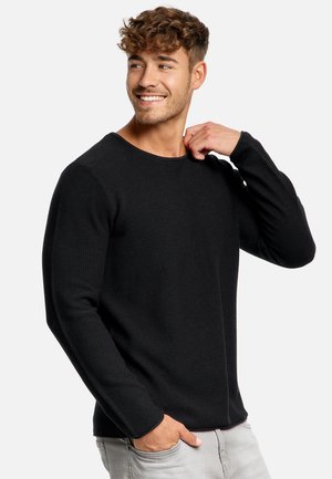 Giovane sorridente con capelli ricci che indossa un maglione nero a maniche lunghe e pantaloni grigio chiaro, con una mano in tasca e l'altra che tocca il colletto.
