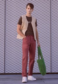 Beige zip vest over a brown t-shirt, burgundy jeans, and white sneakers. Green tote bag. Grated metal background.