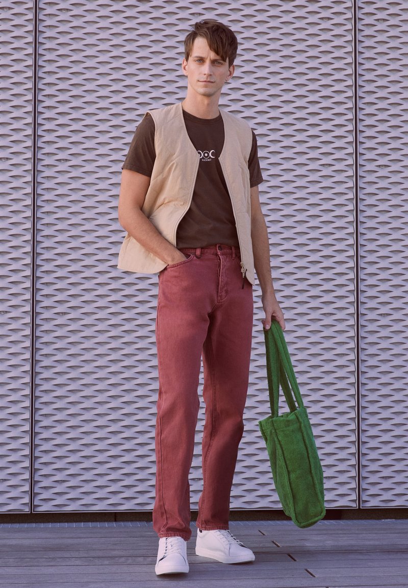Beige zip vest over a brown t-shirt, burgundy jeans, and white sneakers. Green tote bag. Grated metal background.