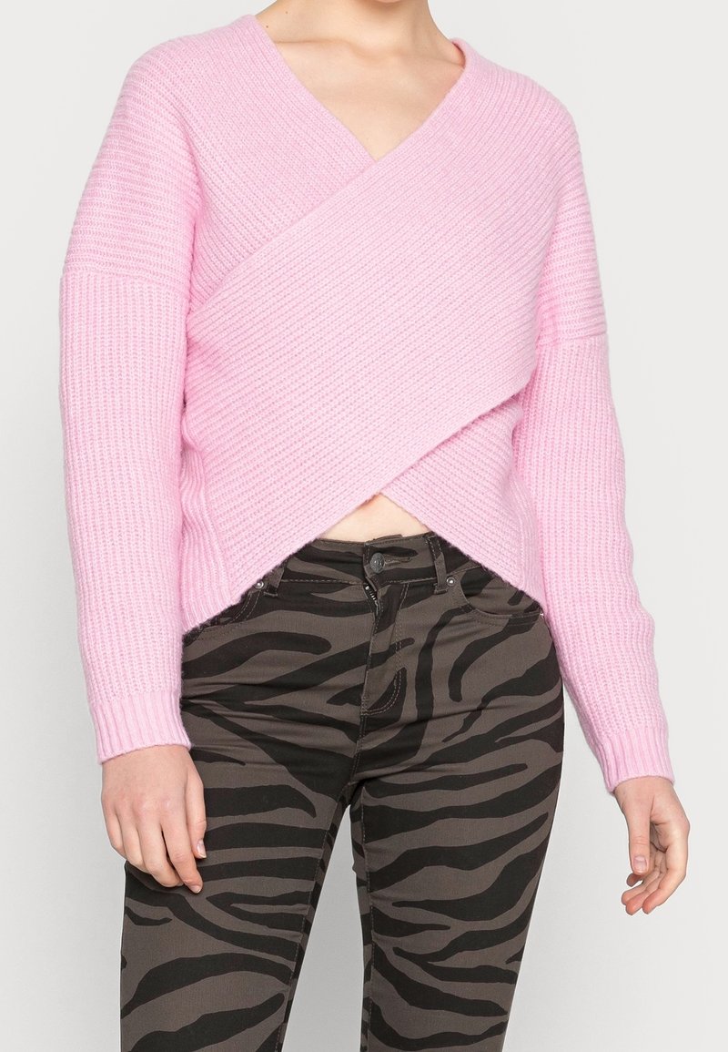 Różowy sweter dziergany o przekładanym kroju i prążkowanej fakturze. W połączeniu z ciemnobrązowymi spodniami z czarnym wzorem zebry.