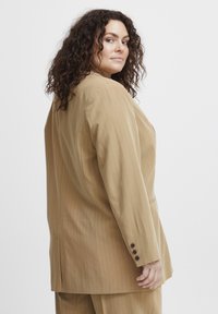 Barna csíkos blézer laza szabással, amelynek hajtókája van, hosszú ujja és háromgombos mandzsettája. Sima anyagtextúra, minimális részletezéssel.