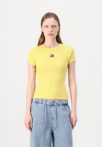 T-shirt jaune à manches courtes et côtelé, présentant un petit logo rectangulaire rouge, blanc et bleu sur la poitrine. Tissu doux et ajusté.