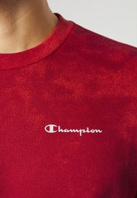 Gros plan sur un t-shirt rouge à col rond avec le logo blanc "Champion" imprimé sur la poitrine, porté par une personne.